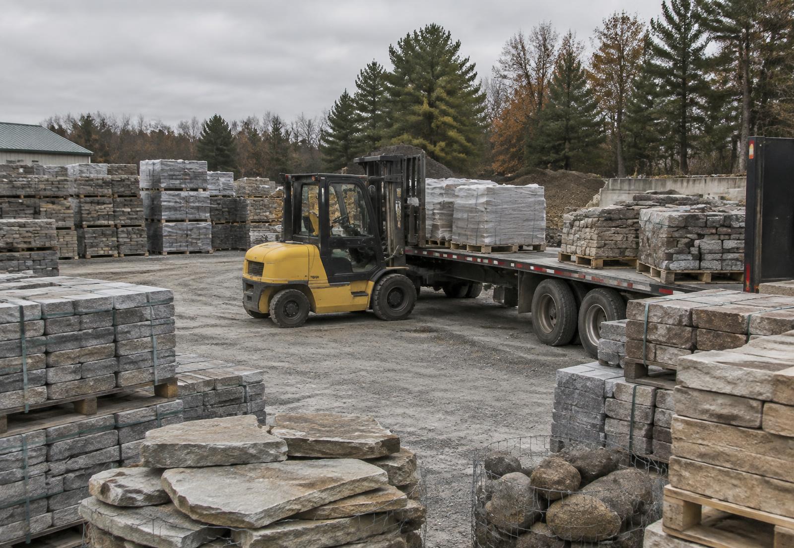Pallets of pavers, wall block and natural stone in a supply yard with a forklift loading a contractor trailer